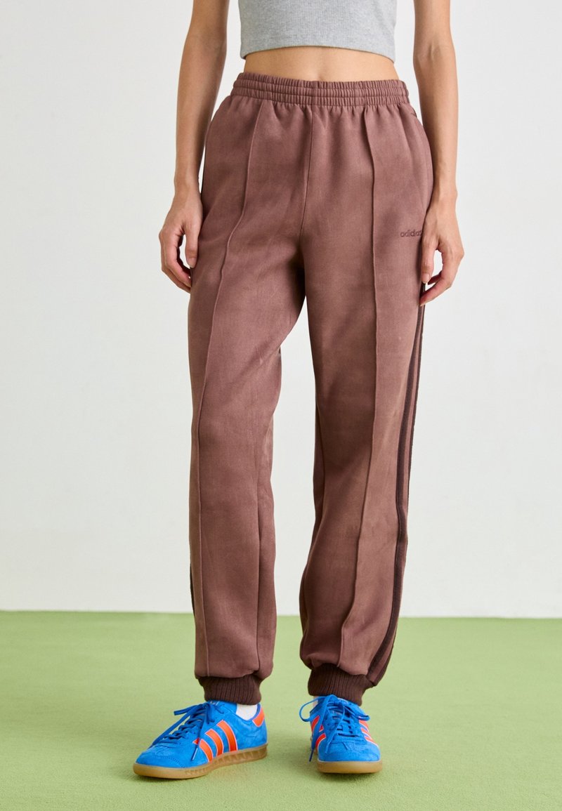 Pantalons de jogging marron avec une finition texturée, taille élastique et rayures latérales. Associés à des chaussures de sport bleues avec des accents orange.