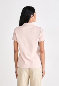 Camisa polo rosa de mangas curtas com colarinho e barra reta, feita de um tecido leve e suave. A vista traseira mostra detalhes de costura mínimos.