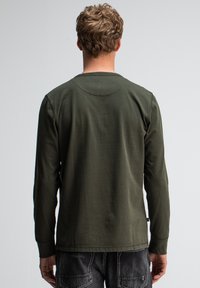 Mann mit kurzen Locken, der ein olivgrünes Langarmshirt und dunkelblaue Jeans trägt, steht vor einer schlichten hellgrauen Wand.