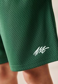 Grüne Sportshorts aus perforiertem Stoff mit einem weißen, gestickten Logo und einem geraden Saum. Nahaufnahme des Materials.