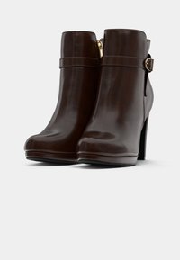 Σκούρες καφέ δερμάτινες μπότες αστραγάλου με φερμουάρ στο πλάι, διακοσμητική ζώνη, λεπτομέρεια με αγκράφα και ψηλές τετράγωνες τακούνια σε λευκό φόντο.