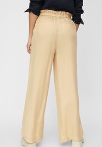 Persona che indossa pantaloni ampi beige con vita elasticizzata, una maglietta nera a maniche lunghe e scarpe bianche, in piedi su uno sfondo semplice.