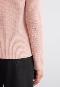 Pull en tricot rose clair avec un motif texturé et des poignets côtelés, associé à un jean sombre, montrant le dos et le détail de la manche gauche.
