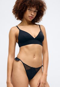 Fekete bralette vékony pántokkal és mély V-nyakkivágással, megfelelő csipkés szélekkel díszített tangával, amely áttetsző pöttyös anyagból készült.