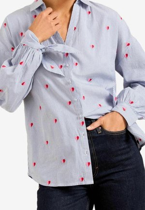 Close-up van een persoon die een blauw-wit gestreept shirt draagt met kleine rode hartjespatronen, de ene hand bij de nek, de andere in een donkere denimzak.