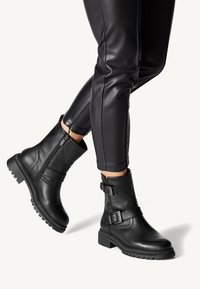 Bottines en cuir noir avec fermeture éclair sur le côté et doubles sangles à boucles, portées avec un pantalon noir ajusté, montrées sur des jambes croisées.