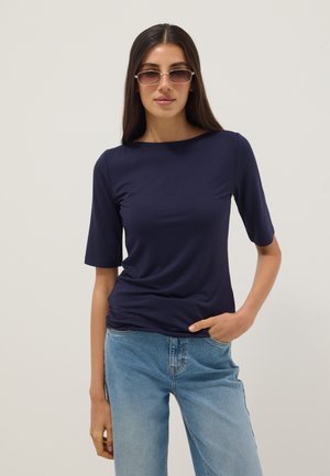 Mujer con blusa azul marino de media manga, jeans azul claro y gafas de sol rectangulares, posando con una mano en el bolsillo sobre un fondo liso.