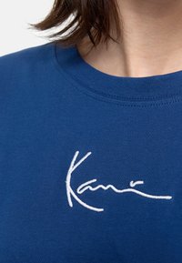 Tums ar jūras zila kokvilnas audumu, uz krūtīm ir balta izšūtā paraksta logo. Apaļa kakla izgriezums ar gludu tekstūru un īsām piedurknēm.