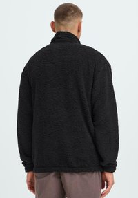 Svart fleece-genser med en teksturert, plysj overflate, høy krage og lange ermer. Løs passform med rett kant.