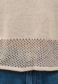 Μπεζ πλεκτό πουλόβερ με ενιαία πάνω περιοχή και ανοιχτό σχέδιο δικτυώματος κοντά στο τελείωμα, πάνω από μπλε τζιν παντελόνι.