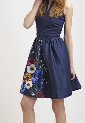 Ärmelloses marineblaues Kleid mit tailliertem Bund und floralem Muster auf einer Seite des ausgestellten Rocks, getragen von einer stehenden Frau.