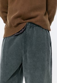 Hnedý rebrovaný sveter navrstvený na tmavosivých kordových nohaviciach so širokými nohavicami a vertikálnymi pruhmi. Viditeľný plochý pás a bočné vrecká.