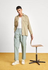 Beige skjorta med knappar över en vit tanktop, ljusgröna byxor och beige sneakers. Trästol bredvid huvudpersonen mot en gul bakgrund.