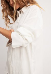 Valkoinen nappipaita kevyestä kankaasta, jossa on kaulus, ylöspäin käännetyt hihat ja hienovarainen tekstuuri. Muotopuoli istuvuus ja etupainikkeet.