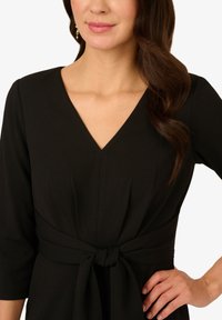 Schwarzes Kleid mit tiefem V-Ausschnitt, Dreiviertelärmeln und einer taillierten Taille, die mit einer Schleife gebunden ist. Glattes Material mit subtilen Texturdetails.