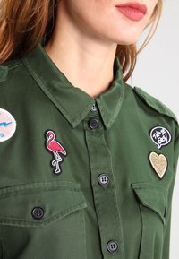 Camicia verde a pulsante con due tasche sul petto, caratterizzata da toppe ricamate: un fenicottero, un cuore e una grafica circolare. Tessuto testurizzato.