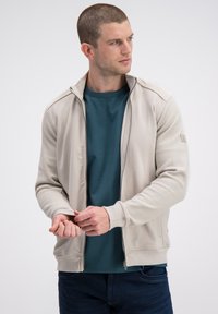 Jasnobeżowa kurtka na zamek z teksturowanymi rękawami, zestawiona z turkusową koszulką z okrągłym dekoltem. Posiada dopasowane mankiety i elegancki design.