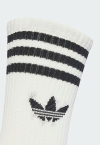 Weißer gestrickter Socken mit einem gerippten Bündchen, das zwei schwarze horizontale Streifen und ein schwarzes Trefoil-Logo aufweist. Zudem hat der Socken eine weiche Textur und ein sportliches Design.
