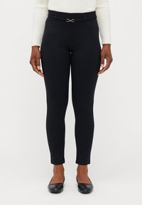 Kvinna i svarta tighta byxor med ett smalt bälte och svarta ballerinan, stående mot en enfärgad vit bakgrund.