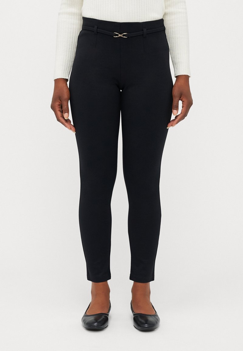 Kvinna i svarta tighta byxor med ett smalt bälte och svarta ballerinan, stående mot en enfärgad vit bakgrund.