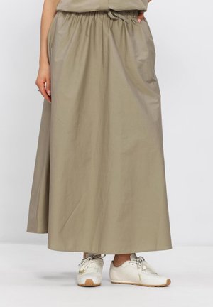 Femme portant une longue jupe beige à taille élastique et des baskets blanches à lacets, debout les mains dans les poches de la jupe.