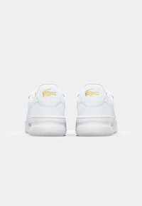 Bílé kožené tenisky s hladkou texturou, zdobené zlatým vyšívaným logem na zadní straně a bílou gumovou podrážkou.