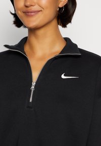 Svart zip-up sweatshirt med hög krage, med en silverdragkedja och en liten vit Nike-logotyp på bröstet. Mjuk tygtextur.