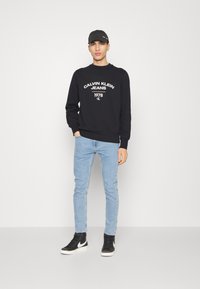 Felpa nera con testo bianco, jeans azzurri chiari e sneaker nere. La felpa presenta un collo a giro e polsini a costine.