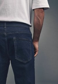 Los jeans de mezclilla azul oscuro cuentan con acentos cosidos, un bolsillo trasero y una cinturilla. La tela tiene una textura suave.