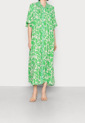 Kobieta w zielono-różowej długiej sukience z liściastym wzorem, zapinanej na guziki z przodu, z krótkimi rękawami, w beżowych sandałach na obcasie z odkrytymi palcami, stojąca na szarej podłodze.