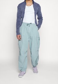 Blå ribbade zip-up kofta, vit tanktop, ljus teal cargobyxor med fickor och lila-rutiga slip-on sneakers.