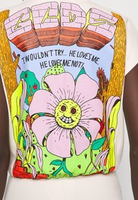 Grafik-T-Shirt mit einer großen, bunten Blume mit einem lächelnden Gesicht, umgeben von lebhaften Pflanzen und Text, auf einem hellen Stoffhintergrund.