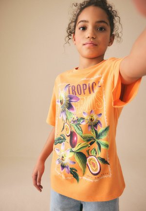 Bambino con maglietta arancione con stampa di fiori tropicali e frutta e jeans azzurri, che tende un braccio in avanti.