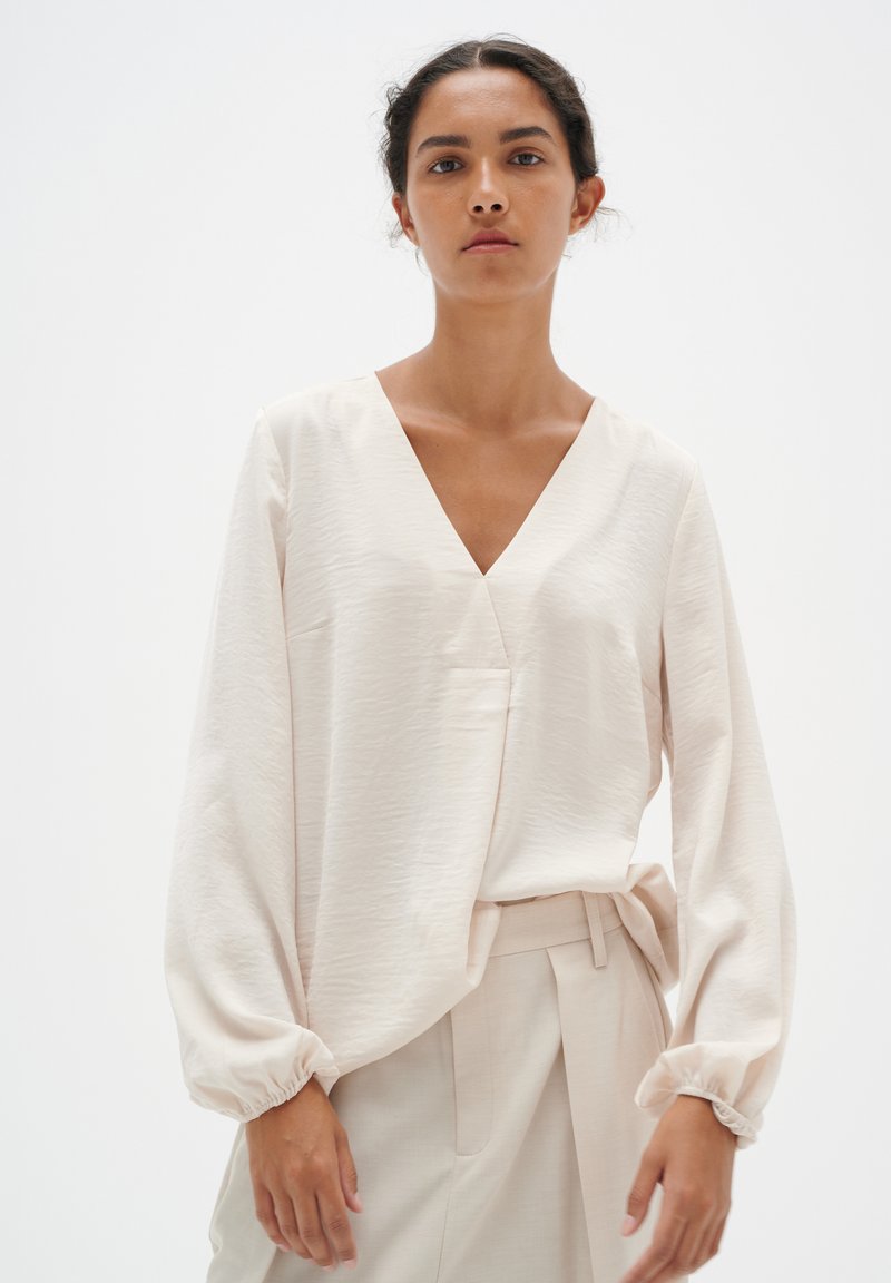 Helle beige Bluse mit langen Ärmeln und tiefem V-Ausschnitt, aus strukturiertem Stoff und mit gerafften Manschetten; kombiniert mit passenden Hosen.