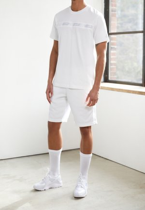 Homme portant une tenue sportive entièrement blanche : chemise à manches courtes, short jusqu'aux genoux, chaussettes montantes et baskets, debout à l'intérieur près d'une fenêtre.