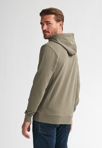 Sudadera verde oliva con cordón y bolsillo tipo canguro, combinada con jeans de mezclilla azules. Tejido suave con un diseño ajustado.
