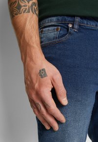 Indigókék farmernadrág, keskeny szabással, réz színű kiegészítőkkel, elülső zsebekkel, és egy kéz pihen a combon, amely egy tetoválást mutat.
