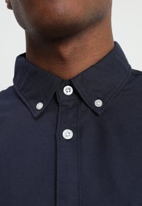 Granatowa koszula z guzikami, z klasycznym kołnierzykiem, dwoma białymi guzikami przy szyi i gładką teksturą materiału.