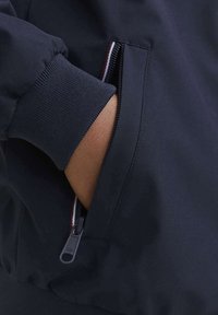 Giacca bomber navy con polsini a coste, dotata di una tasca con zip e una trama di tessuto discreta. Il cursore della zip è nero con un logo sobrio.