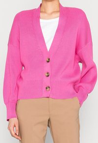 Kvinna som bär en ljusrosa ribbad kofta med tre bruna knappar över en vit topp och beige byxor, med handen i fickan på en enfärgad bakgrund.