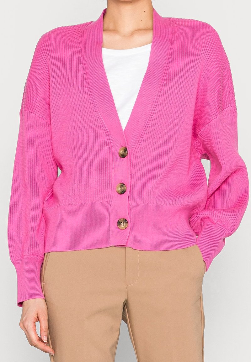 Kvinna som bär en ljusrosa ribbad kofta med tre bruna knappar över en vit topp och beige byxor, med handen i fickan på en enfärgad bakgrund.