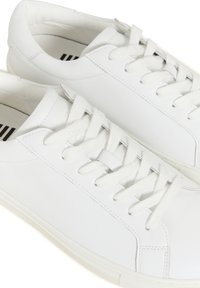 Paire de baskets basses blanches en cuir avec lacets blancs et semelles en caoutchouc, vues de près sous un angle supérieur.