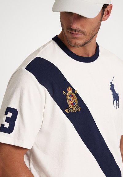 Μπλουζάκι από βαμβάκι σε κρεμ χρώμα με ναυτικό μπλε διαγώνια ρίγα. Διαθέτει κεντημένο λογότυπο και τον αριθμό "3" στο μανίκι. Ριμπ λαιμόκοψη.