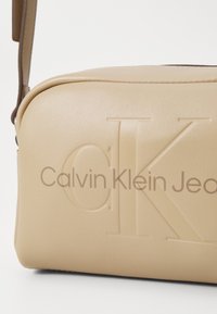 Borsa in ecopelle beige con logo "CK" in rilievo e testo "Calvin Klein Jeans". Forma quadrata, texture liscia, accentuata da una tracolla marrone.