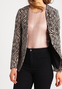 Blazer à imprimé zèbre noir et beige à coupe ample, associé à un pull côtelé rose clair. Un pantalon noir complète la tenue.