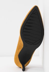 Chaussure à talons hauts jaune moutarde avec un bout pointu et une semelle en caoutchouc noire présentant une texture nervurée. Taille 37 marquée sur la semelle.