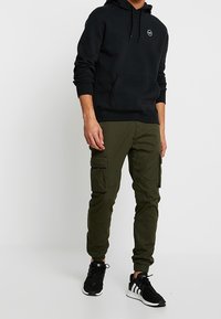 Sudadera con capucha negra y bolsillo frontal, combinada con pantalones cargo verdes que tienen bolsillos laterales. Usada con zapatillas deportivas en blanco y negro.