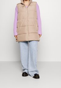 Beige dunvest med sidelommer og høj krave, båret over en lys lilla ribstrikket sweater og lys blå vide jeans. Sorte platform sko.