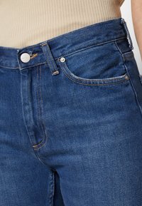 Dark blue denim jeans with a high waist, featuring contrasting orange stitching, a metal button closure, and classic five-pocket design.