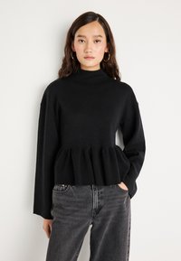 Svart strikkegenser med høy hals, lange ermer og peplumkant, parret med grå jeans med en myk tekstur.